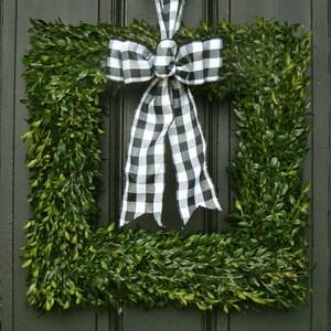 May include: A black and white checkered bow is tied to a green boxwood wreath. The wreath is shaped like a square and is made of boxwood branches.