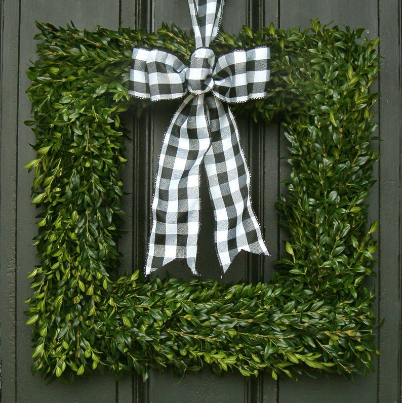 May include: A black and white checkered bow is tied to a green boxwood wreath. The wreath is shaped like a square and is made of boxwood branches.