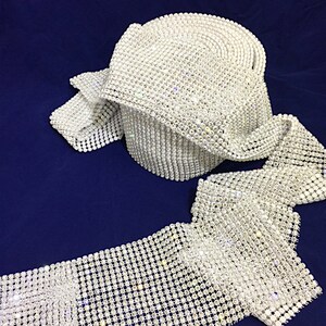 May include: A roll of white rhinestone trim, with a clear, sparkly finish. The trim is made of small, round rhinestones that are sewn together in a grid pattern.