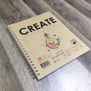 May include: A spiral-bound sketchbook with the word "CREATE" in large black letters on the cover. The cover is tan with a watercolor illustration of a lightbulb. The sketchbook is 8.5 x 11 inches and contains 50 pages.