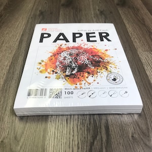 May include: A pack of 100 sheets of multi-media paper. The paper is medium-heavy weight and has a semi-smooth finish. The pack is wrapped in plastic and has a barcode and QR code on the back. The front of the pack features a colorful abstract design with a leopard print.