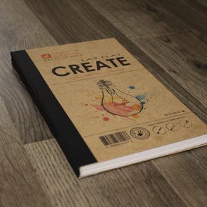 May include: A notebook with a kraft paper cover and black spine, featuring the word "CREATE" in bold letters. The cover also displays a lightbulb illustration with watercolor accents and the text "LAY FLAT".