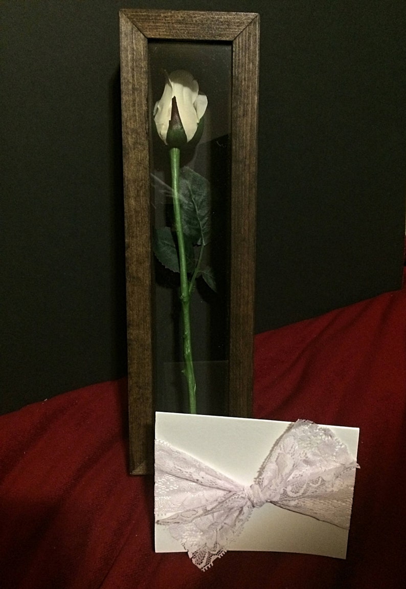 May include: A white rose in a wooden shadow box. The rose has a green stem and leaves. A white card with a pink lace bow is in front of the shadow box. The background is a dark red fabric and black.