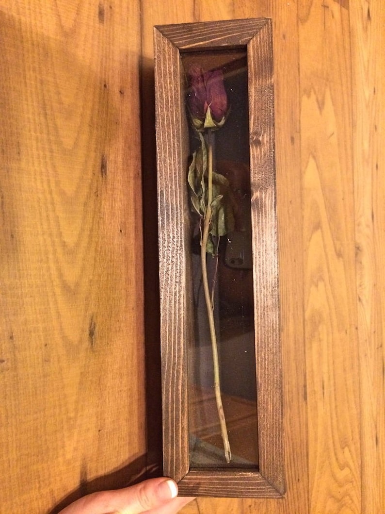 May include: A framed dried rose in a rectangular wooden shadow box. The rose is a deep burgundy color with green leaves and stem. The wooden frame has a dark brown finish and a glass front.