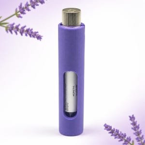 May include: A purple perfume bottle with a gold cap is encased in a matching purple holder. The bottle has a rectangular window revealing the white label with the word "HERETIC". Lavender sprigs are in the background.