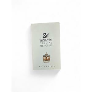May include: A rectangular white product box featuring the black Swarovski swan logo, the text SWAROVSKI CRYSTAL MEMORIES, and a small graphic of a gold and clear crystal carousel. The word CLASSICS is printed in black at the bottom of the box.
