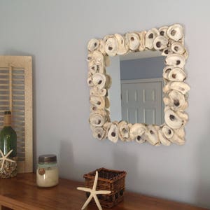 May include: Square mirror with a frame made of oyster shells. The mirror hangs on a light blue wall above a wooden surface. Other coastal decor includes a green bottle, a small basket, and a starfish.