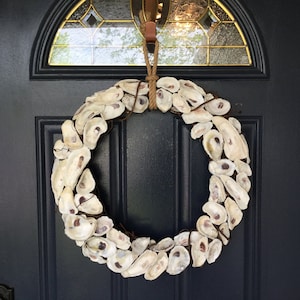 May include: A circular wreath constructed from numerous off-white oyster shells arranged in a dense, overlapping pattern. The shells are secured to a circular frame with dark brown wire and the wreath hangs from a thick, tan jute rope loop.