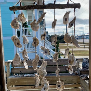 May include: A rustic wooden dowel with oyster shells hanging from it. The shells are strung on twine and are of various sizes and shapes. The dowel is hanging from the ceiling of a porch.