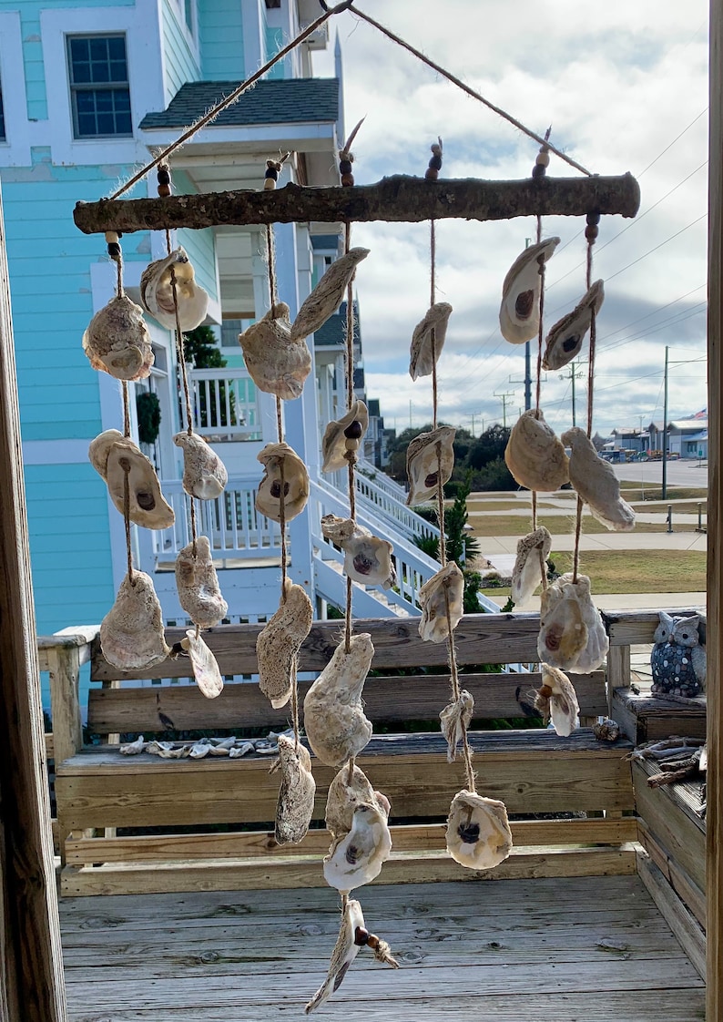 May include: A rustic wooden dowel with oyster shells hanging from it. The shells are strung on twine and are of various sizes and shapes. The dowel is hanging from the ceiling of a porch.