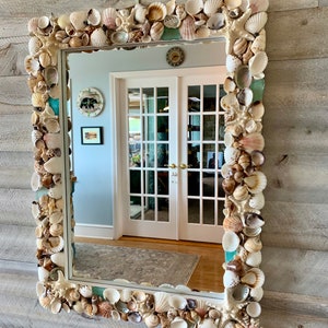 May include: A rectangular mirror with a decorative frame made of seashells. The frame is made of a variety of seashells in different shapes, sizes, and colors. The mirror is reflecting a room with a door and a rug.
