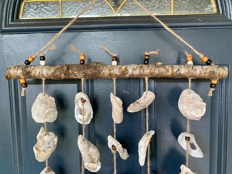 May include: A rustic wooden branch with hanging oyster shells and brown and black beads. The branch is suspended from a twine string and the shells are arranged in a symmetrical pattern.