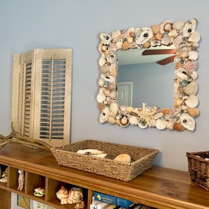 May include: Square mirror with a frame decorated with various seashells. The mirror is mounted on a light blue wall. A wooden shelf below holds decorative items, including a woven basket and a driftwood piece.