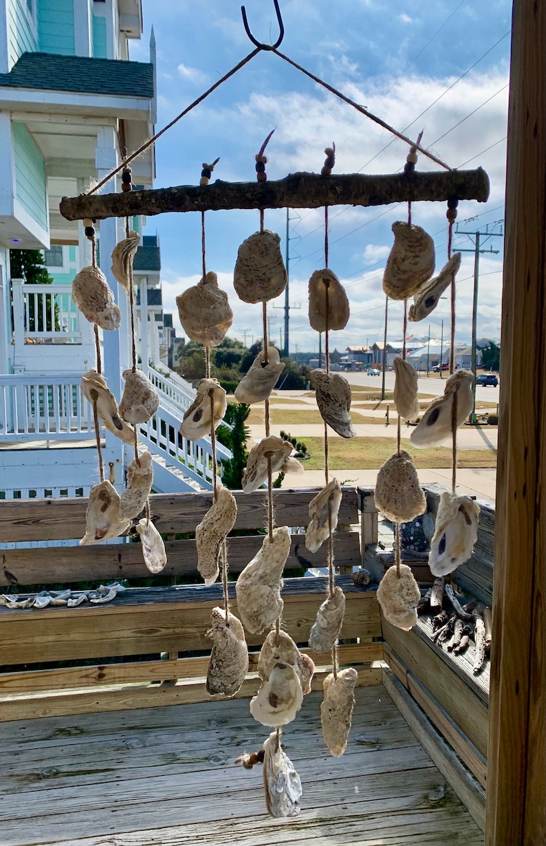 May include: A rustic oyster shell wind chime hangs from a wooden branch. The shells are strung on twine and are arranged in a cascading pattern. The wind chime is hanging from a hook on a wooden porch.