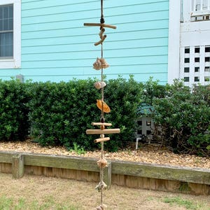 May include: A seashell and driftwood mobile hanging from a hook. The mobile features a variety of seashells and driftwood pieces strung together with twine. The mobile is hanging in front of a light blue wall.