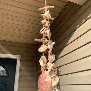 May include: A seashell wind chime with various sized and colored seashells hanging from wooden dowels and twine. The chime is hanging from a hook on a light brown wooden surface.