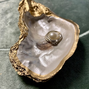 May include: A white oyster shell ring dish with a gold rim. A pearl ring with a clear stone setting is resting in the dish.
