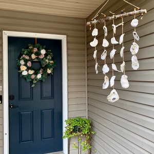 May include: A white door with a pink and green floral wreath hanging on it. A wooden branch with oyster shells hanging from it is on the right side of the door. A green plant is in a pot on a metal stand.