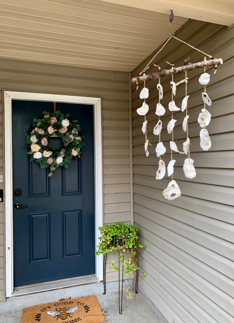 May include: A white door with a pink and green floral wreath hanging on it. A wooden branch with oyster shells hanging from it is on the right side of the door. A green plant is in a pot on a metal stand.