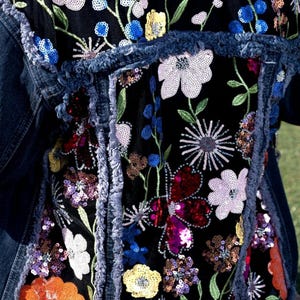 May include: A denim jacket with a black mesh panel featuring colorful embroidered flowers and sequins. The flowers are in shades of pink, red, blue, yellow, and orange.