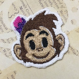 Könnte beinhalten: Ein brauner gestickter Affen-Patch mit weißem Rand und einer lila und pinkfarbenen Krone. Der Affe hat ein braunes Gesicht mit weißen Augen und einem weißen Lächeln.