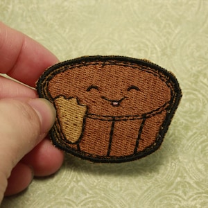 以下が含まれることがあります： 茶色のフェルト生地のパッチに、笑顔の漫画のカップケーキが描かれています。カップケーキは茶色のフェルト生地のアイシングと黒いフェルト生地の輪郭で描かれており、茶色のフェルト生地の棒を持っています。