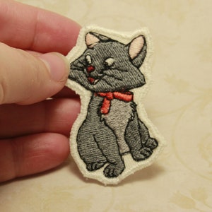 以下が含まれることがあります： 赤いリボンをつけた灰色の猫の刺繍ワッペン。猫は目を閉じて少し微笑んで座っています。ワッペンはクリーム色の縁取りで、手で持たれています。