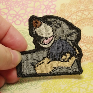 Op de afbeelding: Geborduurde patch met een cartoon beer karakter in grijs, bruin en beige. De patch heeft een zwarte rand en wordt door een hand vastgehouden. De beer lacht met zijn mond open. De patch is waarschijnlijk voor kleding of accessoires.