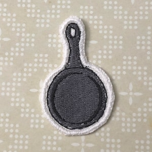 Op de afbeelding: Een wit geborduurd pleister van een zwarte koekenpan met een handvat.