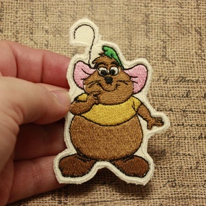 以下が含まれることがあります： 茶色のネズミの刺繍パッチ。黄色のシャツ、緑色の帽子、ピンク色の耳をしています。ネズミは微笑んで、顎に手を当てています。