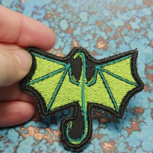 以下が含まれることがあります： 緑と黒の刺繍が施されたアイロン接着パッチ。翼を広げたドラゴンが描かれています。