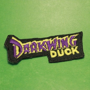 May include: A black embroidered patch with the words "Darkwing Duck" in yellow and purple lettering.