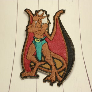 May include: An embroidered patch featuring a brown, anthropomorphic dragon with red wings. The dragon is standing with its arms crossed and has a white mane. The patch is approximately 4 inches tall.