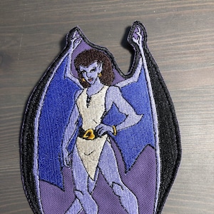 May include: An embroidered patch featuring a purple gargoyle with wings outstretched. The gargoyle is wearing a white tunic and has brown hair and blue eyes.