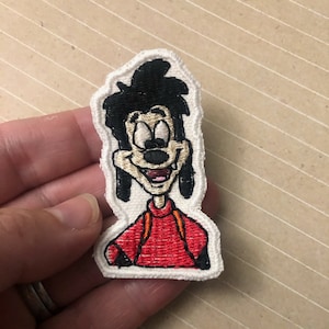 Può includere: Un toppa ricamata da stirare con un personaggio dei cartoni animati con i capelli neri, una camicia rossa e un contorno bianco. Il personaggio ha un grande sorriso e indossa uno zaino.