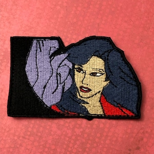 Könnte beinhalten: Gestickter Aufnäher mit einer Comic-Frau mit langen, dunkelblauen Haaren, die eine rote Jacke trägt und zur Seite schaut. Die Frau ist teilweise von einer lila Hand verdeckt.