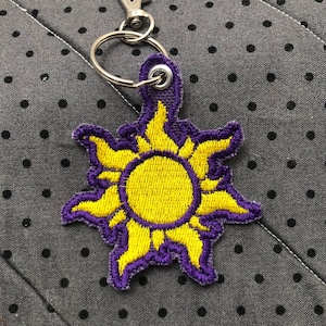 May include: A yellow sun with purple rays, embroidered on a fabric keychain. The keychain has a silver ring and a silver clip.