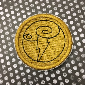 May include: Embroidered patch with a gold circular design featuring a black lightning bolt and a stylized swirl. The patch has a gold border and a white backing. The background is a gray surface with white polka dots.