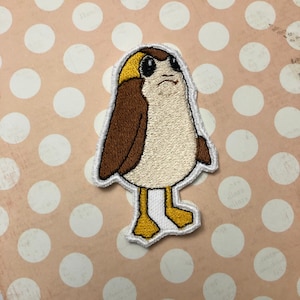 Op de afbeelding: Een geborduurde strijk-op patch met een cartoonfiguur met een bruin lichaam, een wit hoofd en een gele snavel. Het figuur heeft grote ogen en een kleine mond. De patch is omgeven door een witte rand.