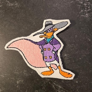May include: Embroidered patch featuring Darkwing Duck in a purple coat, grey hat, and pink cape. The cartoon duck has an orange beak and feet, with a teal scarf. The patch is outlined in white.