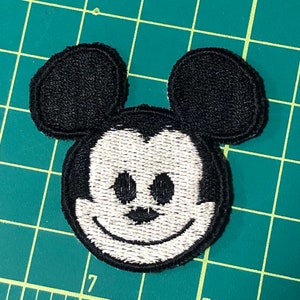 Peut inclure: Patch brodé noir et blanc représentant le visage de Mickey Mouse avec de grandes oreilles.