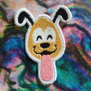 Op de afbeelding: Geborduurde strijkpatch van een cartoonhond met een geel gezicht, witte ogen, een zwarte neus en een roze tong die uitsteekt. De hond heeft zwarte oren en een witte omtrek.