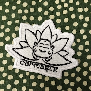May include: Embroidered patch featuring a smiling cartoon figure within a lotus flower design. The patch is white with black outlines and text that reads "carmaste". The patch is set against a green fabric with white polka dots.