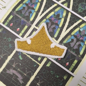 Puede incluir: Un parche bordado de corona de color dorado con costuras blancas. El parche está sobre una superficie blanca con una ventana de vitrales en el fondo.