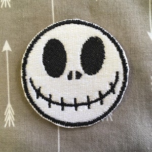 May include: White embroidered patch with a black outline, featuring a smiling Jack Skellington face from the Nightmare Before Christmas.