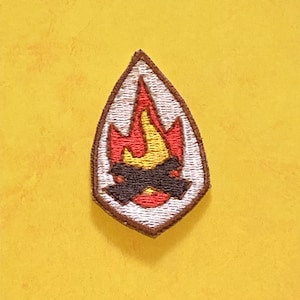 Op de afbeelding: Een bruin en wit geborduurde patch met een geel en rood vlamontwerp. De vlam bevindt zich in een zwarte vorm die lijkt op een kampvuur.