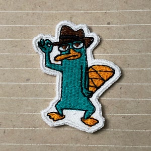 May include: Embroidered iron-on patch featuring a cartoon platypus wearing a brown fedora and sunglasses. The platypus is teal blue with an orange tail fin.