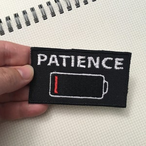 May include: Black rectangular patch with the word "PATIENCE" embroidered in white above a black battery icon with a red bar indicating low power. The patch is held against a grid-lined notebook.
