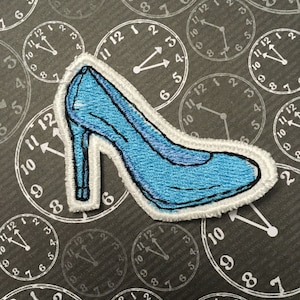 Peut inclure: Écusson brodé d'un escarpin bleu clair avec une bordure blanche. Le patch est posé sur un fond d'horloges avec des cadrans blancs et des fonds noirs.