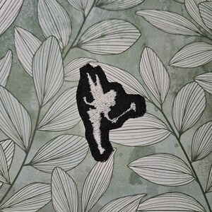 May include: A black embroidered patch featuring a white silhouette of a fairy. The patch is set against a background of green and white leaves. The fairy is depicted in a dynamic pose, with outstretched arms and a flowing skirt. The patch is likely for clothing or accessories.